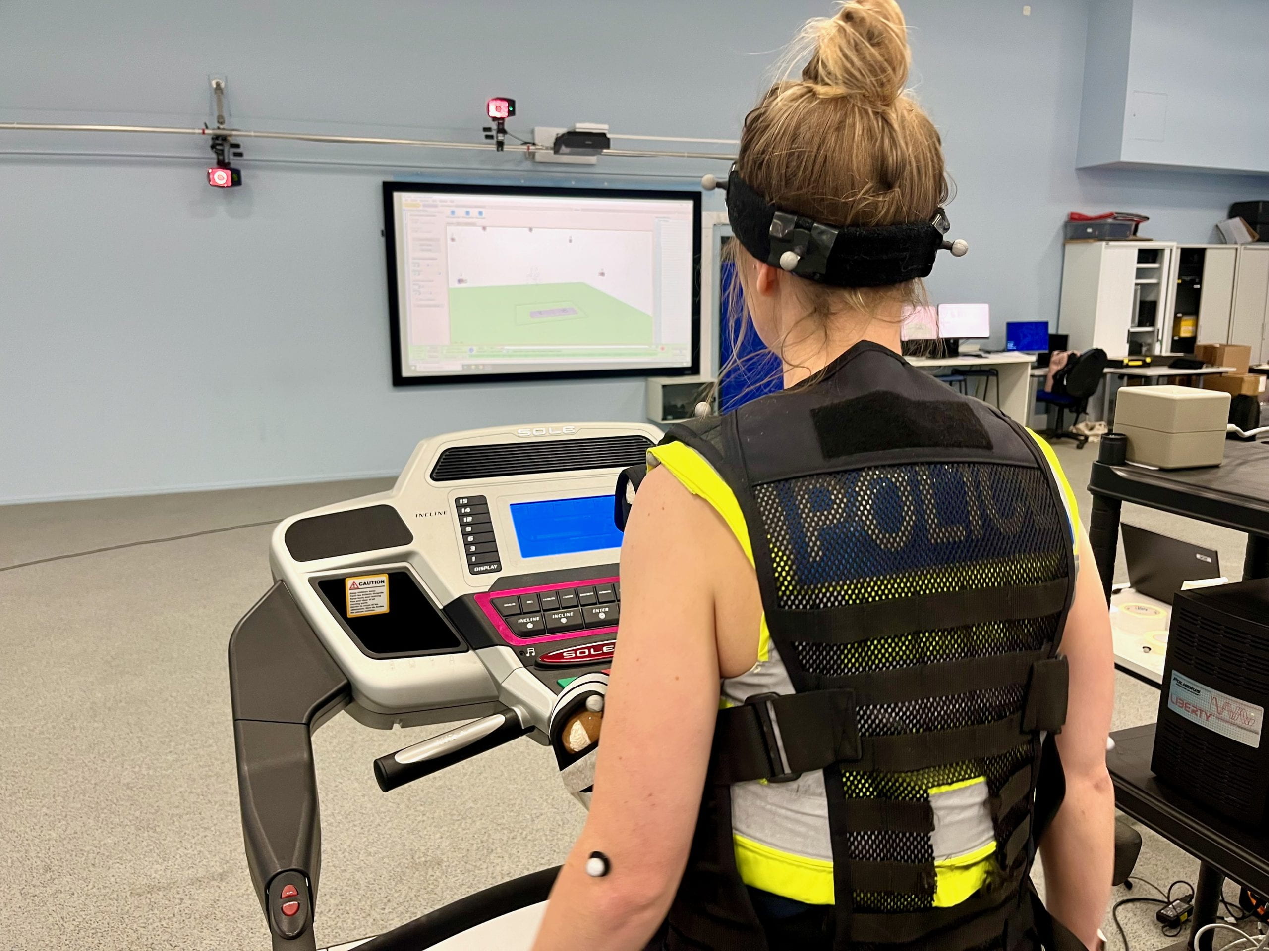 A woman wearing a police vest walks on a treadmill in a motion capture laboratory