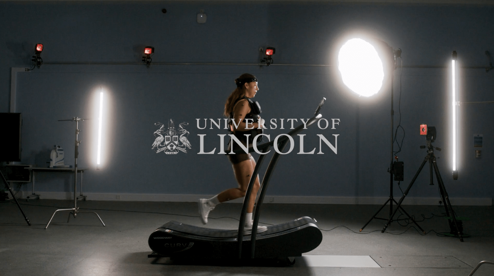 A woman in a tactical vest runs on a treadmill in a motion capture laboratory.